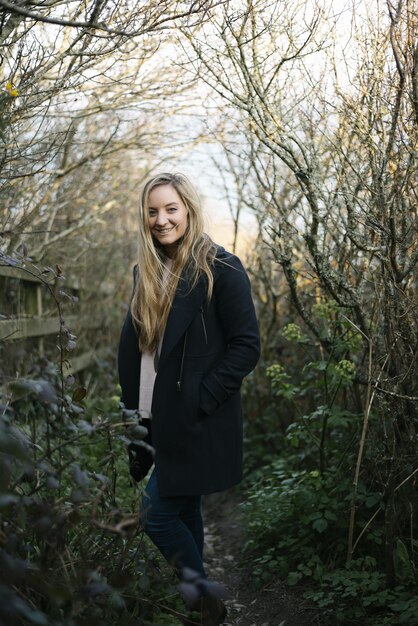Joven mujer rubia con un abrigo negro de pie sobre un camino rodeado de árboles sin hojas