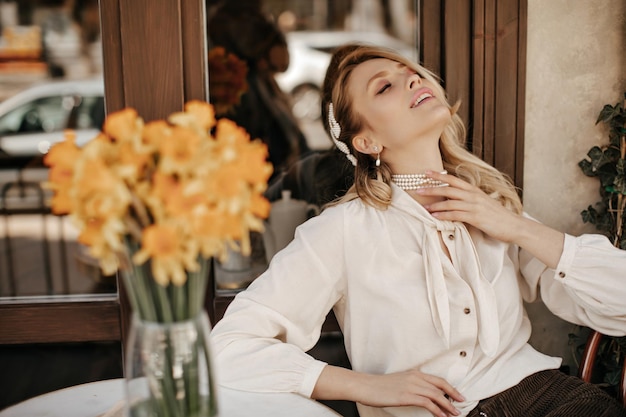 Joven mujer rubia alegre con collar de perlas, blusa elegante blanca, toca suavemente las poses del cuello con los ojos cerrados y sonríe en el café de la calle