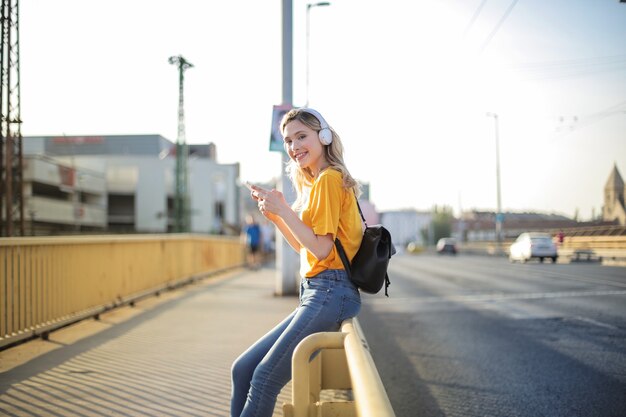 Joven mujer rubia apoyada en una valla de puente y enviando mensajes de texto en su teléfono mientras escucha música