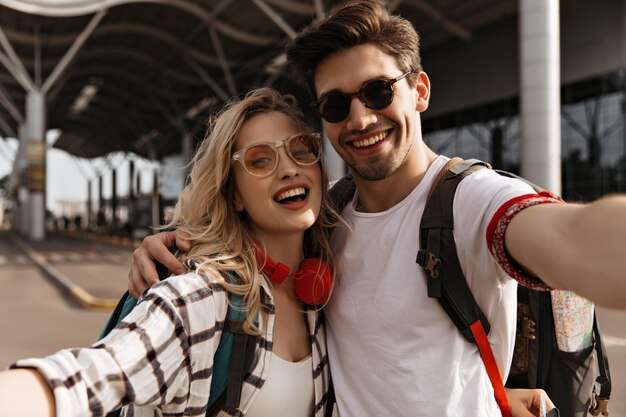 Joven mujer rubia atractiva con camisa a cuadros y elegante hombre moreno con gafas de sol sonríe y se toma selfie Retrato de un par de viajeros cerca del aeropuerto