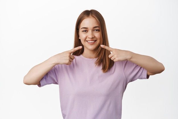 Joven mujer rubia en camiseta apuntando a sus dientes blancos, boca, de pie contra la pared blanca con gran sonrisa feliz