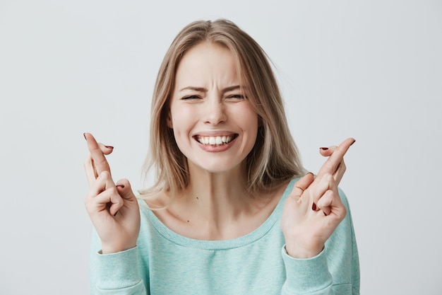 Joven mujer rubia caucásica cerrando los ojos cruzando los dedos con esperanza, anticipando noticias importantes. Hermosa mujer con expresión esperanzada gesticulando con los dedos, mostrando dientes blancos