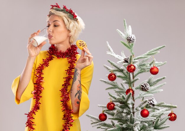 Joven mujer rubia con corona de Navidad y guirnalda de oropel alrededor del cuello de pie cerca del árbol de Navidad decorado con vaso de leche y galletas bebiendo leche aislado sobre fondo blanco