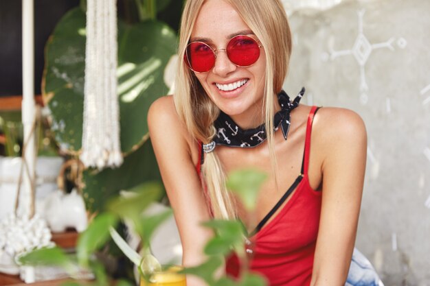 Joven mujer rubia con elegantes gafas de sol rojas posando en un café