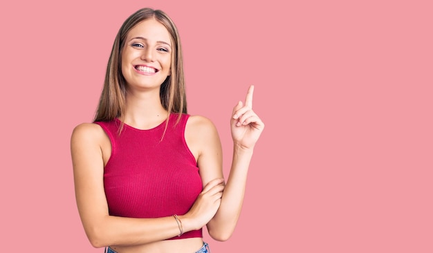 Joven mujer rubia hermosa con una elegante camisa de verano con una gran sonrisa en la cara, señalando con la mano y el dedo hacia un lado mirando la cámara.