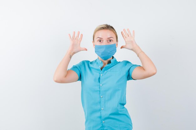 Joven mujer rubia mostrando gesto de rendición en ropa casual, máscara y mirando asustado