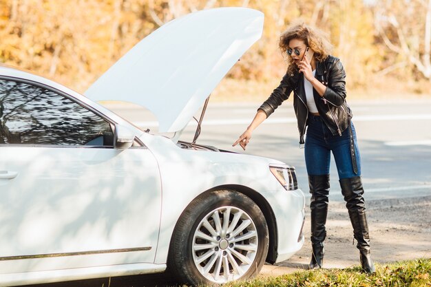 Joven mujer rubia de pie cerca de un coche roto con capota abierta hablando por su teléfono móvil mientras espera ayuda.