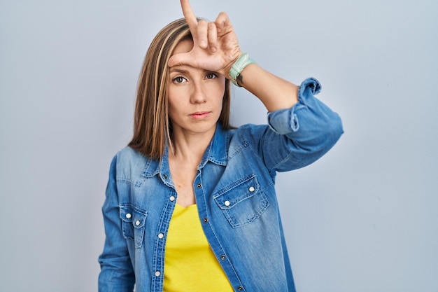 Foto gratuita joven mujer rubia de pie sobre fondo azul burlándose de la gente con los dedos en la frente haciendo gestos perdedores burlándose e insultando