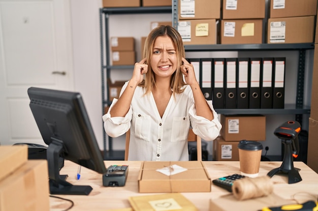 Joven mujer rubia que trabaja en el comercio electrónico de pequeñas empresas cubriendo las orejas con los dedos con una expresión molesta por el ruido de la música a todo volumen. concepto sordo.