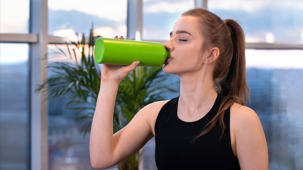 Joven mujer rubia en ropa deportiva está bebiendo agua durante un entrenamiento con los ojos cerrados
