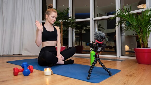 Joven mujer rubia en ropa deportiva sobre una estera de yoga saludando a la cámara de video de grabación frente a ella