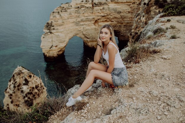 Joven mujer rubia sentada en un acantilado y disfrutar de la vista del océano y la playa