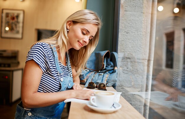 Joven mujer rubia sentada en la cafetería y leyendo