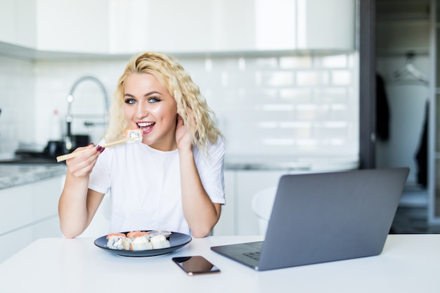 Joven mujer rubia sentada en su cocina comer sushi en casa mesa trabajando con una computadora portátil en la mañana