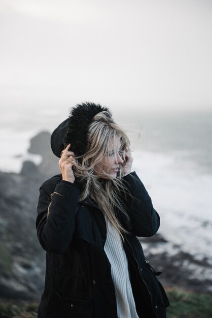 Joven mujer rubia con sombrero caminando sobre una costa rocosa del mar en un clima ventoso