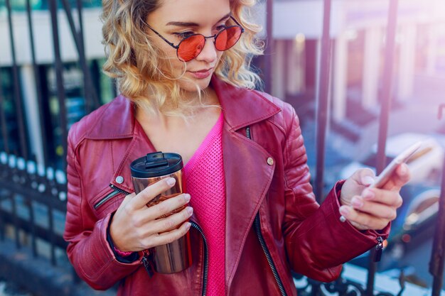 Joven mujer rubia soñadora caminando en la ciudad y utilizando teléfonos inteligentes. Cerrar detalles. Elegante chica moderna con café. Vellos ventosos.