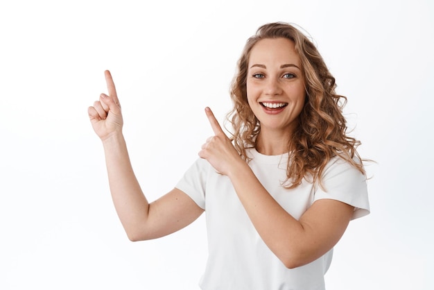 Joven mujer rubia sonriendo alegre señalando el espacio de copia de la esquina superior izquierda que muestra un anuncio de pie sobre fondo blanco