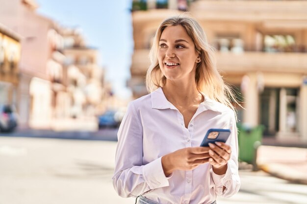 Joven mujer rubia sonriendo confiada usando un teléfono inteligente en la calle