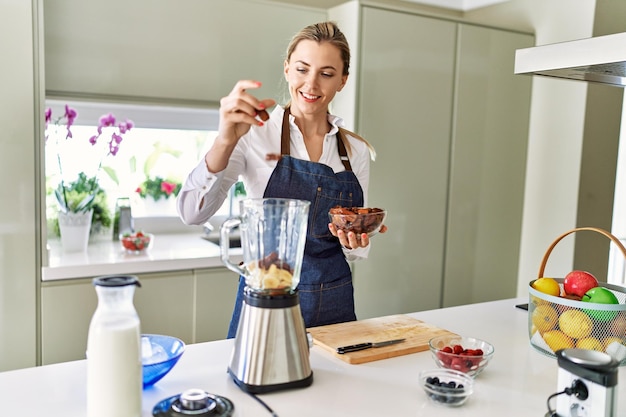 Joven mujer rubia sonriendo confiada vertiendo fechas en la licuadora en la cocina