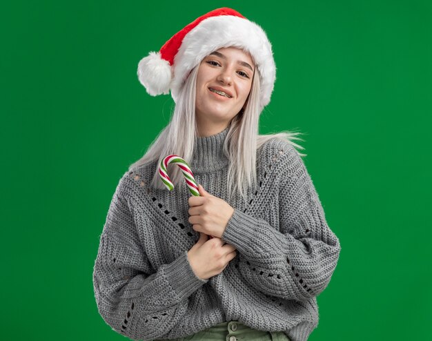 Joven mujer rubia en suéter de invierno y gorro de Papá Noel con bastón de caramelo mirando a la cámara sonriendo feliz y positivo de pie sobre fondo verde