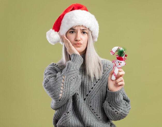 Joven mujer rubia en suéter de invierno y gorro de Papá Noel con bastón de caramelo de Navidad confundido de pie sobre la pared verde