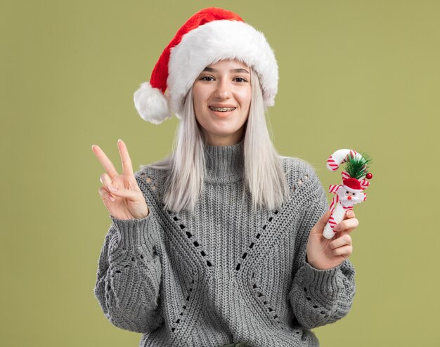 Joven mujer rubia en suéter de invierno y gorro de Papá Noel con bastón de caramelo de Navidad sonriendo alegremente mostrando v-sign parado sobre la pared verde