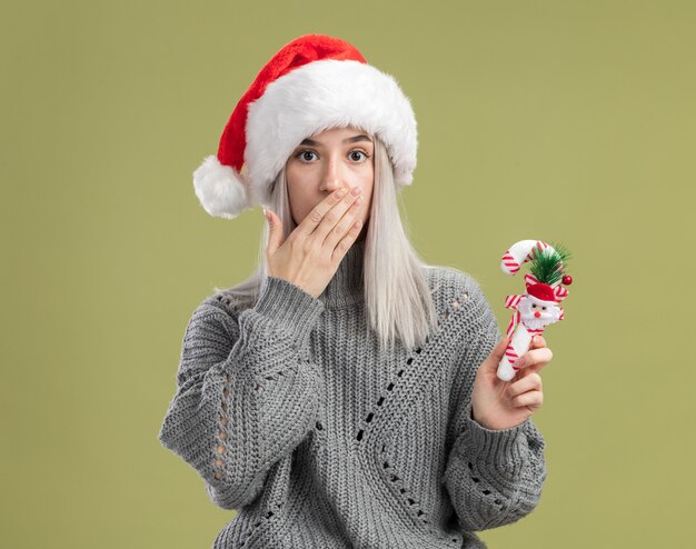Joven mujer rubia en suéter de invierno y gorro de Papá Noel con bastón de caramelo de Navidad sorprendido cubriendo la boca con la mano de pie sobre la pared verde
