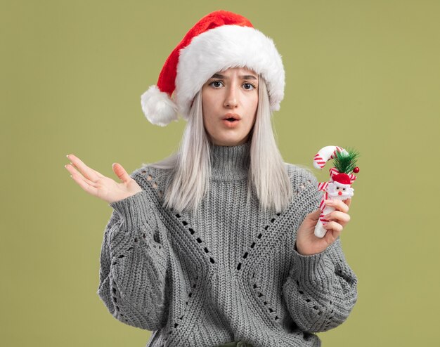 Joven mujer rubia en suéter de invierno y gorro de Papá Noel con bastón de caramelo de Navidad sorprendido de pie sobre la pared verde