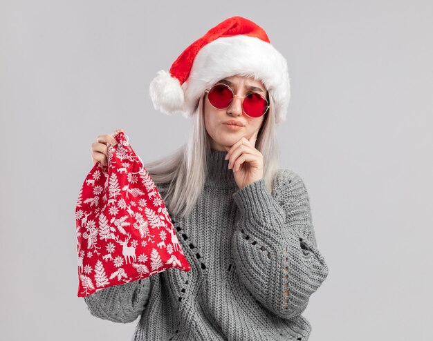 Joven mujer rubia en suéter de invierno y gorro de Papá Noel con bolsa roja de santa con regalos de Navidad mirando a un lado desconcertado