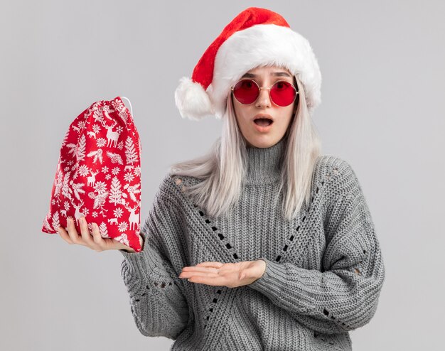 Joven mujer rubia en suéter de invierno y gorro de Papá Noel con bolsa roja de santa con regalos de Navidad presentando con el brazo de la mano mirando sorprendido de pie sobre la pared blanca