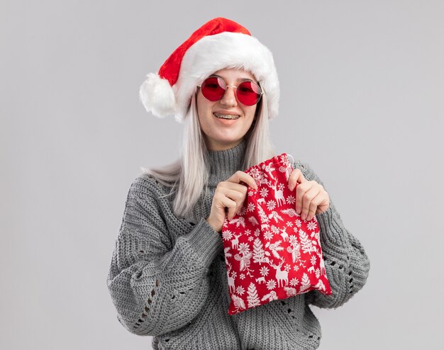 Joven mujer rubia en suéter de invierno y gorro de Papá Noel con bolsa roja de santa con regalos de Navidad con una sonrisa en la cara de pie sobre la pared blanca