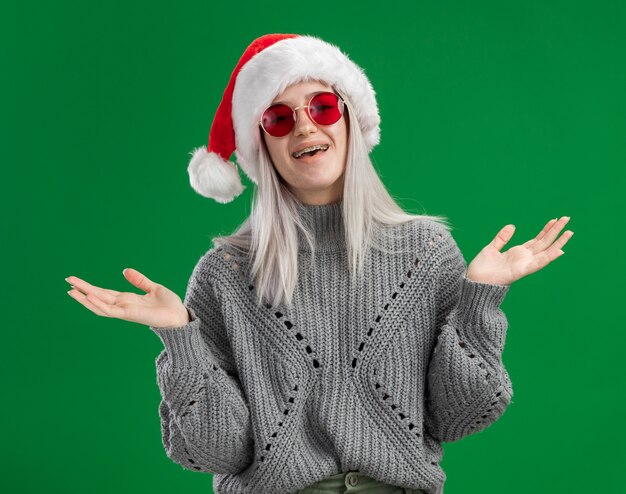 Joven mujer rubia en suéter de invierno y gorro de Papá Noel con gafas rojas mirando a la cámara feliz y positivo sonriendo alegremente de pie sobre fondo verde