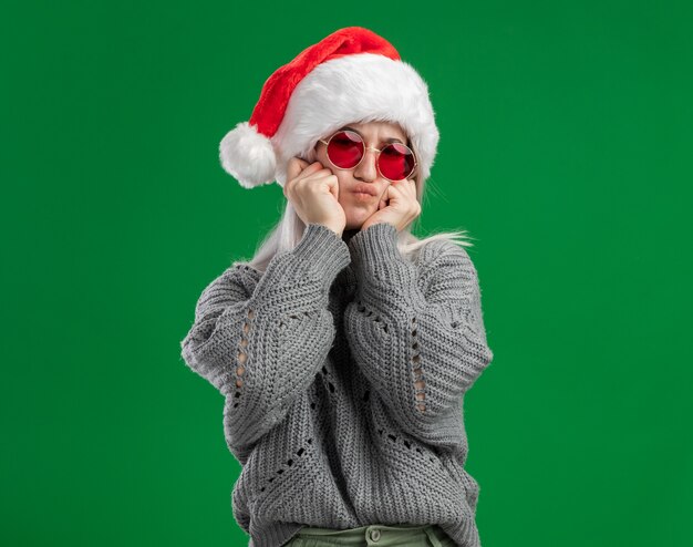 Joven mujer rubia en suéter de invierno y gorro de Papá Noel con gafas rojas mirando a la cámara haciendo muecas divirtiéndose de pie sobre fondo verde