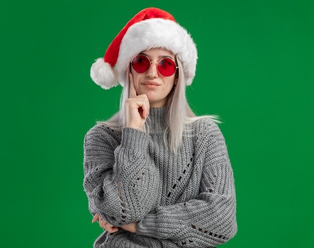 Joven mujer rubia en suéter de invierno y gorro de Papá Noel con gafas rojas mirando a un lado con expresión pensativa pensando de pie sobre fondo verde