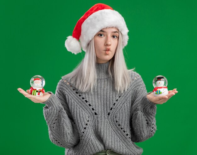 Joven mujer rubia en suéter de invierno y gorro de Papá Noel con globos de nieve de juguete de Navidad mirando a la cámara confundido teniendo dudas sobre fondo verde