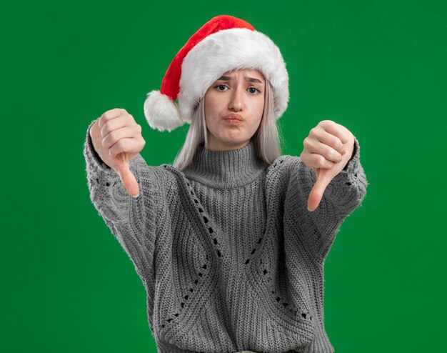 Joven mujer rubia en suéter de invierno y gorro de Papá Noel mirando a la cámara disgustado mostrando los pulgares hacia abajo de pie sobre fondo verde