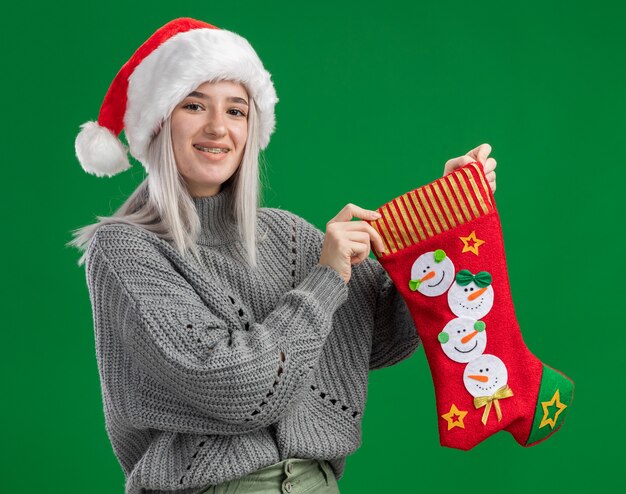 Joven mujer rubia en suéter de invierno y gorro de Papá Noel sosteniendo calcetines de Navidad mirando a la cámara sonriendo alegremente feliz y positivo de pie sobre fondo verde