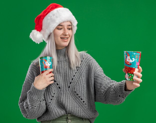 Joven mujer rubia en suéter de invierno y gorro de Papá Noel sosteniendo coloridos vasos de papel mirando la taza sonriendo con cara feliz de pie sobre fondo verde