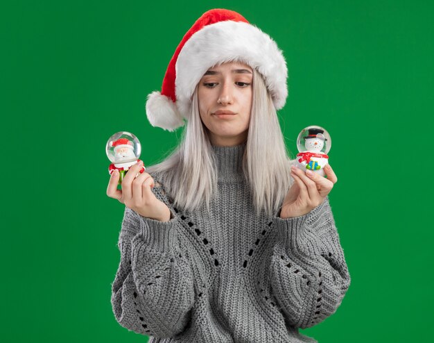 Joven mujer rubia en suéter de invierno y gorro de Papá Noel sosteniendo globos de nieve de juguete de Navidad mirando a la cámara confundido tratando de tomar una decisión de pie sobre fondo verde