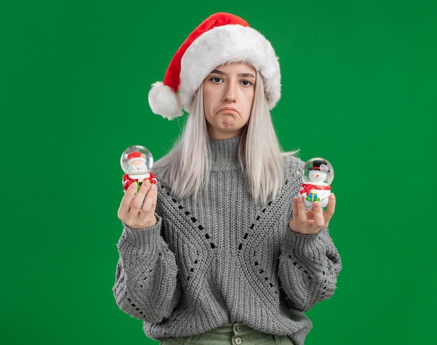 Joven mujer rubia en suéter de invierno y gorro de Papá Noel sosteniendo globos de nieve de juguete de Navidad mirando a la cámara con expresión triste frunciendo los labios sobre fondo verde