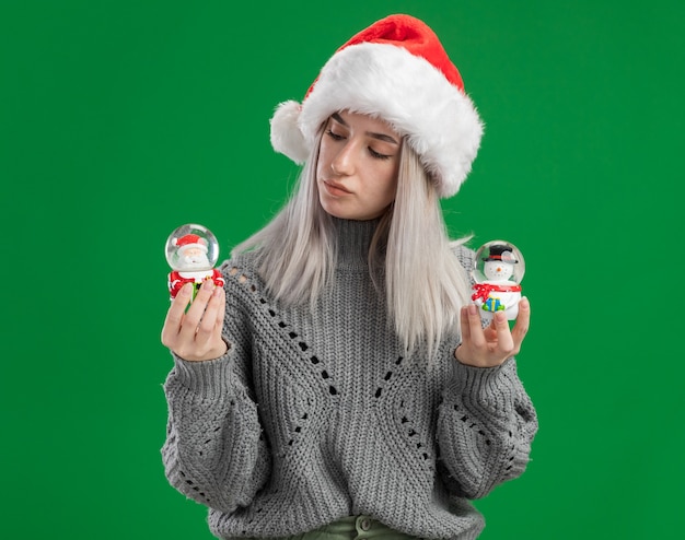 Foto gratuita joven mujer rubia en suéter de invierno y gorro de papá noel sosteniendo globos de nieve de juguete de navidad mirando intrigado de pie sobre fondo verde