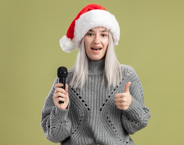 Joven mujer rubia en suéter de invierno y gorro de Papá Noel sosteniendo el micrófono con una sonrisa en la cara mostrando los pulgares para arriba sobre la pared verde