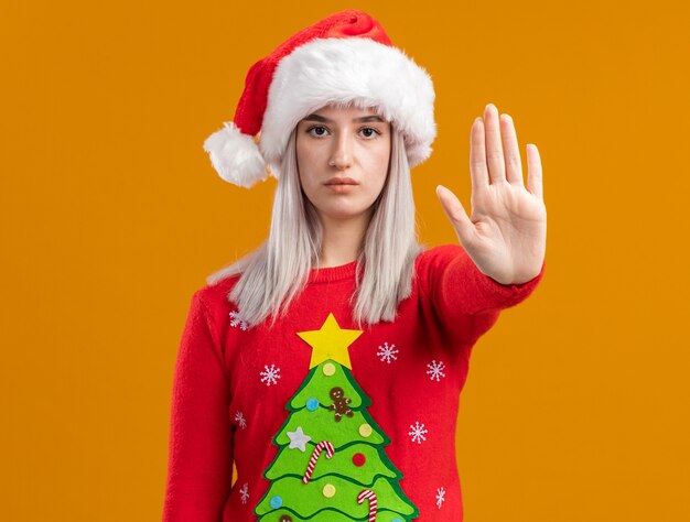 Joven mujer rubia en suéter de Navidad y gorro de Papá Noel con cara seria haciendo gesto de parada con la mano de pie sobre la pared naranja