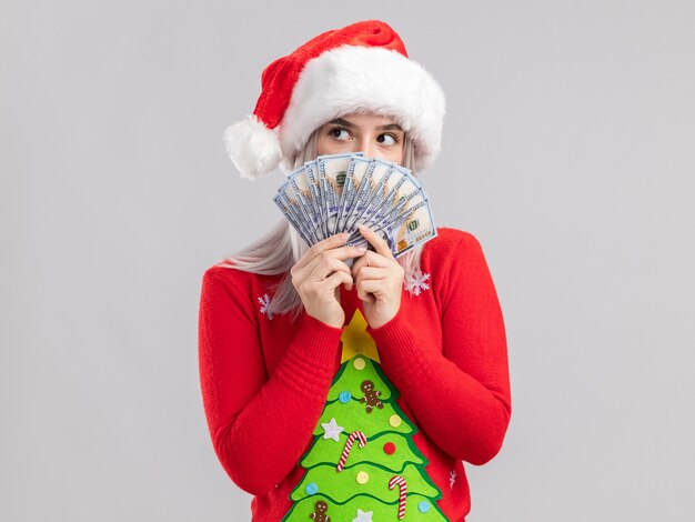 Joven mujer rubia en suéter de Navidad y gorro de Papá Noel con efectivo mirando a un lado con cara seria