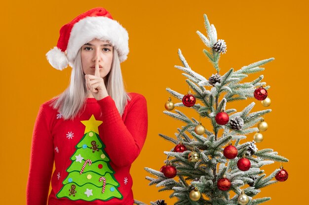 Joven mujer rubia en suéter de Navidad y gorro de Papá Noel haciendo gesto de silencio con el dedo en los labios