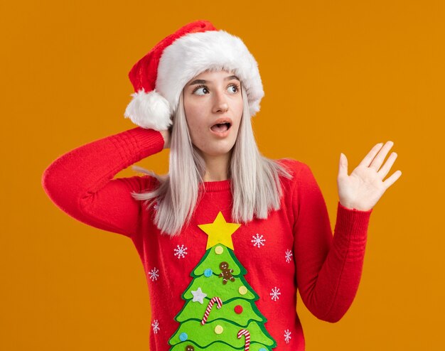 Joven mujer rubia en suéter de navidad y gorro de Papá Noel mirando asie asombrado con el brazo levantado de pie sobre fondo naranja