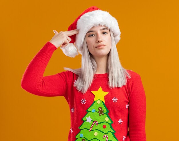 Joven mujer rubia en suéter de Navidad y gorro de Papá Noel mirando a la cámara cansado y aburrido haciendo un gesto de pistola sobre el templo de pie sobre fondo naranja