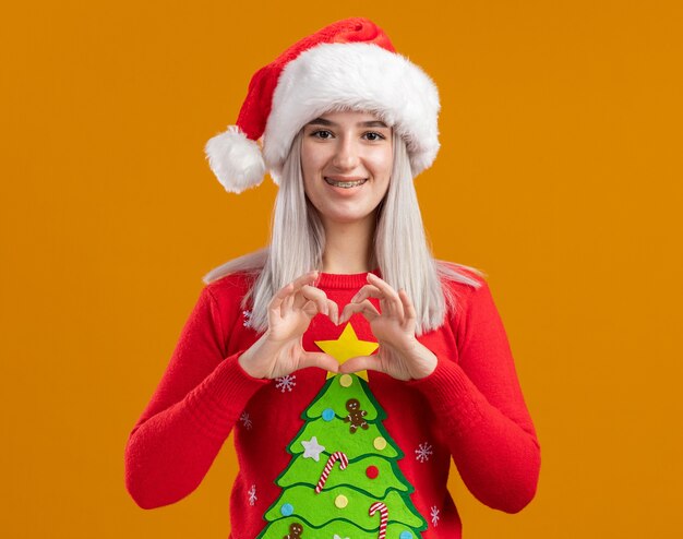 Joven mujer rubia en suéter de Navidad y gorro de Papá Noel mirando a la cámara con una sonrisa en la cara haciendo gesto de corazón con los dedos de pie sobre fondo naranja