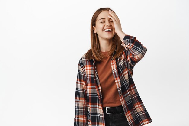 Joven mujer rubia toca su rostro, riendo y sonriendo, expresión de cara feliz, pose despreocupada, de pie con camisa casual a cuadros y jeans negros, pared blanca