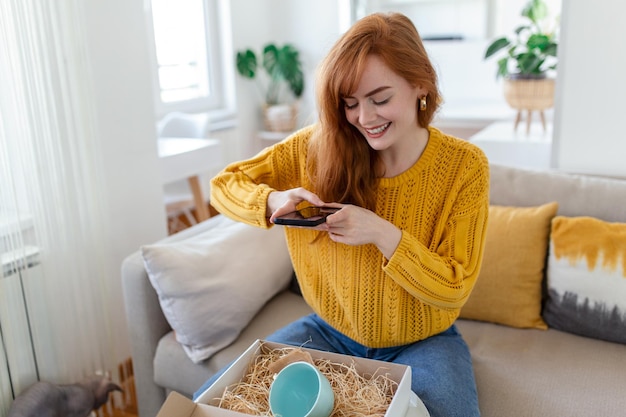 Joven mujer satisfecha feliz cliente adicta a las compras sentada en el sofá desempacando la caja de entrega de paquetes concepto de envío de compras en línea tomando fotos del producto para publicar en las redes sociales